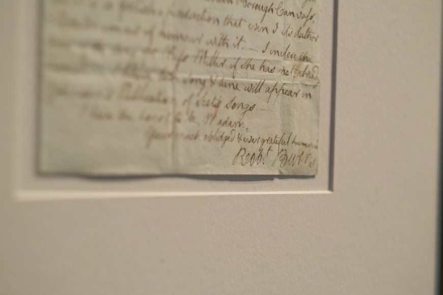 A close-up photo of the end of a handwritten letter from Robert Burns, where he has signed his name on the bottom right corner. Burns has used a brownish ink on parchment-style paper. The letter has been mounted on cream card.