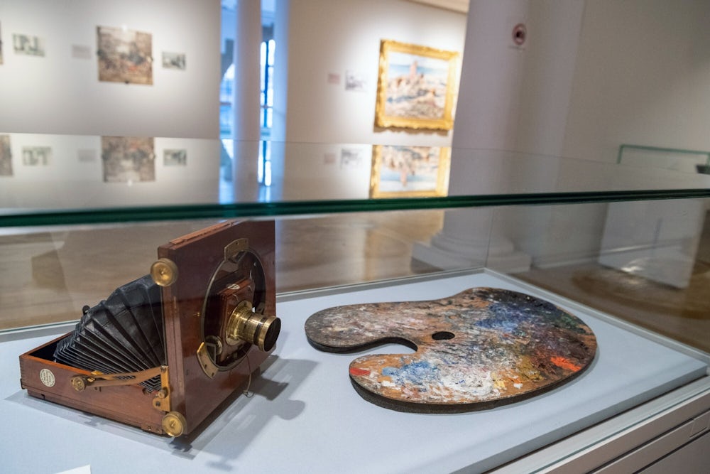 A glass-topped case containing an old-fashioned, folding box camera made of wood with brass fittings and black leather bellows. On the right is a kidney-shaped painter’s palette covered in paint.