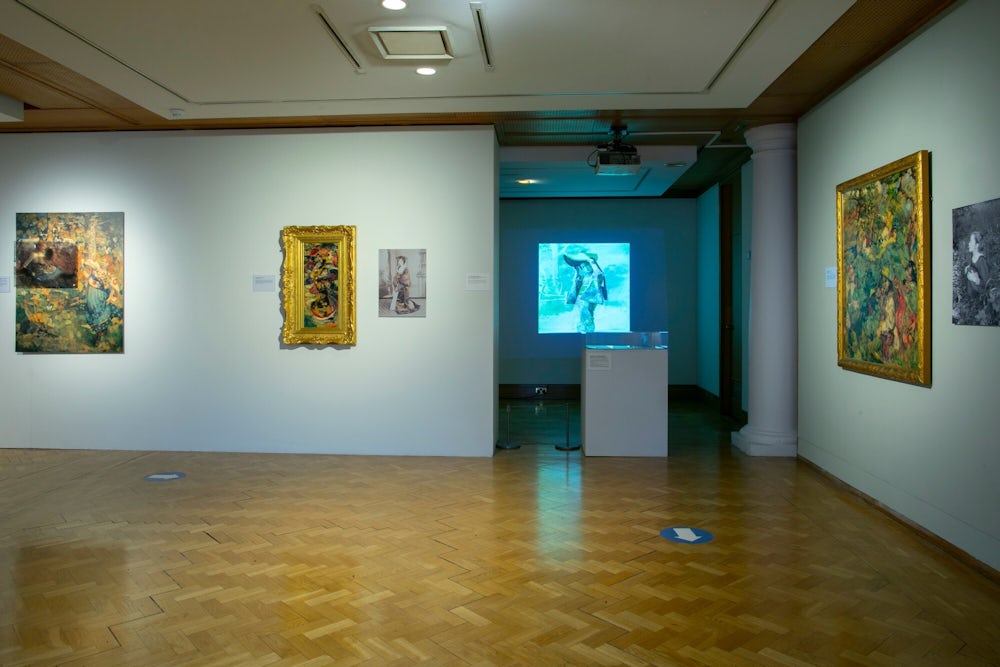 A gallery space with white walls and wooden parquet floors. Three paintings are visible on the walls, and a projected image of a photo is visible in the background. In front of the projected image is a glass-topped case.