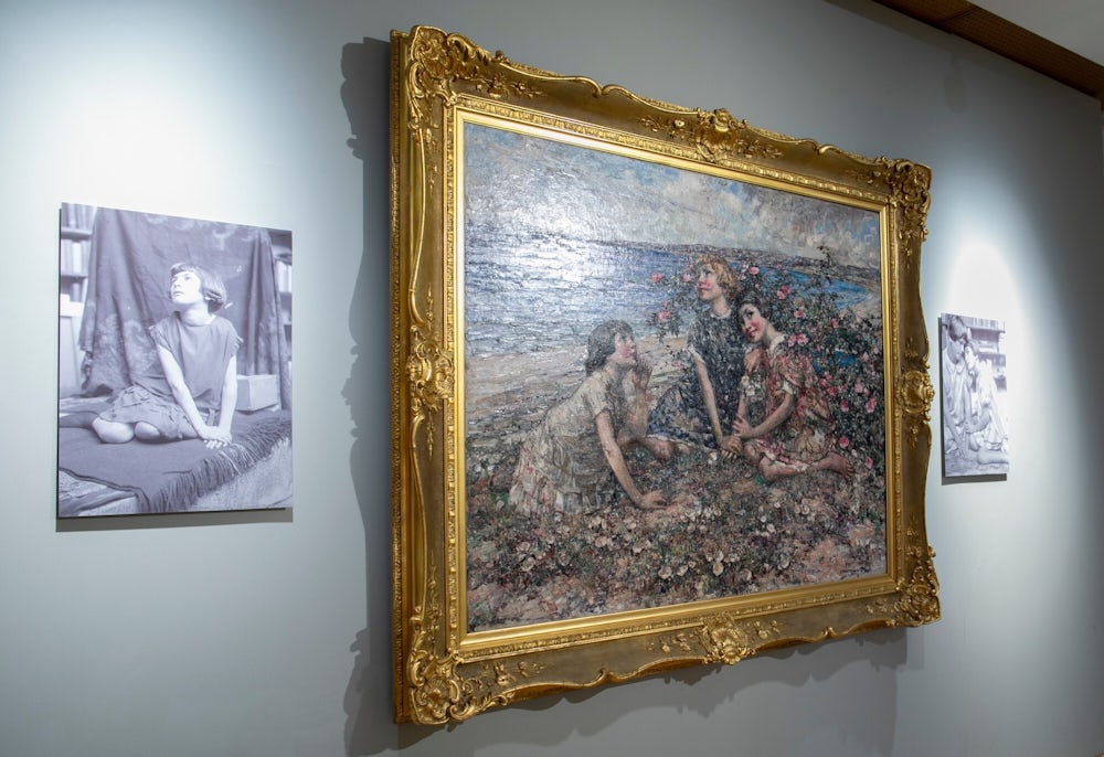 A white wall with a painting in a gilt frame. The painting shows three girls sitting down surrounded by flowers. Behind them a bay with the sea and a sandy beach is visible. Either side of the painting are reproductions of two black and white photos of girls in the same positions as the girls in the painting.